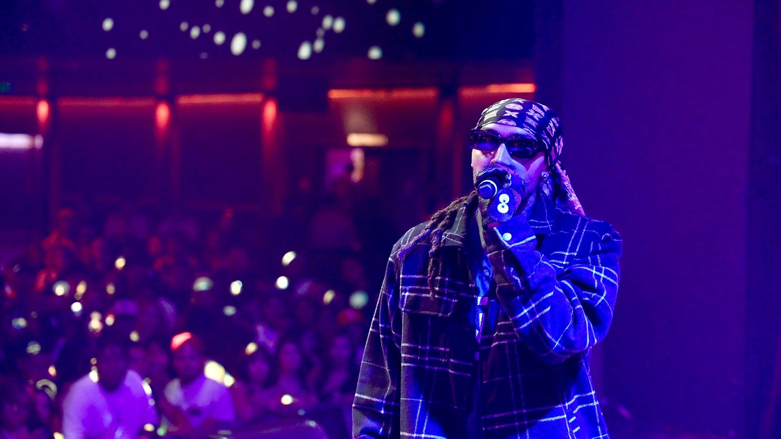 Kilohana in sunglasses and a plaid jacket rapping on stage at The Novo under blue and purple light, phone lights in the crowd behind him